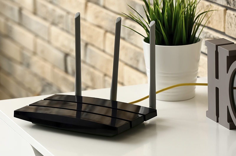 Schwarzer WLAN-Router mit drei Antennen auf Tisch neben Pflanze im Büro